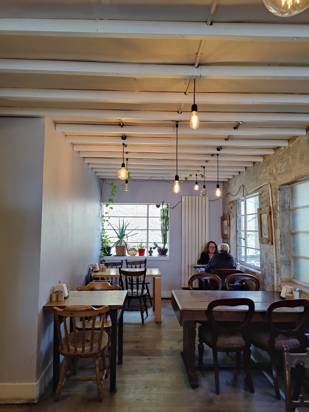 The long room of the cafe with wooden tables, bentwood chairs and a row of hanging bulbs running to a bright window